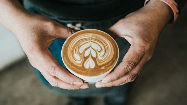 A person holding a latte gently with both hands. The latte art looks like a series of connected hearts. 
