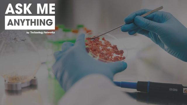 Scientist in blue gloves examines cultured meat in a petri dish using a metal tool, with lab equipment in the background. 