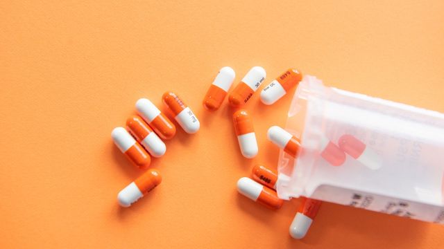 A clear prescription bottle spills white and orange pills onto an orange background. 