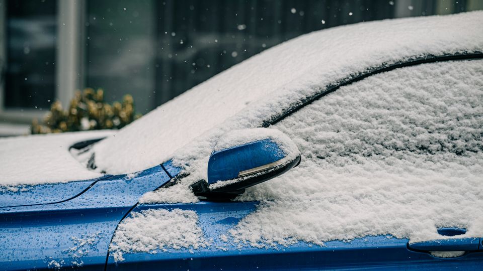 A close-up photograph of a blue car's hood, windows and front door, covered under a thin layer of frost and snow, against a snowy city backdrop.