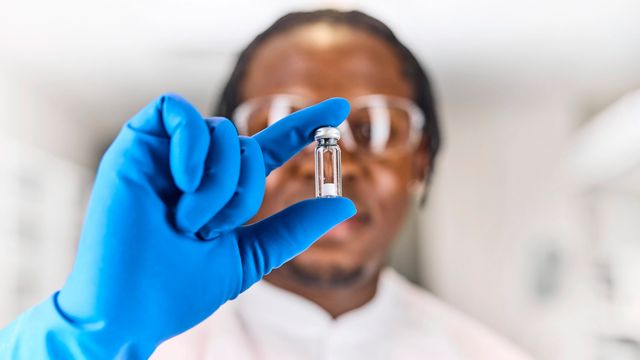 Scientist wearing blue gloves and safety glasses holds up a small vial in a laboratory setting 