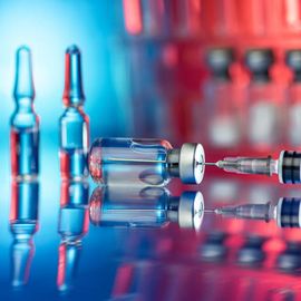 Close-up of vaccine vials and a syringe with reflections on a glossy surface under blue and red light 